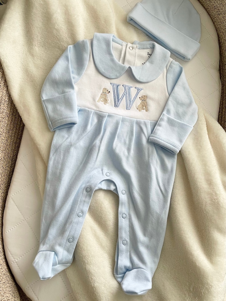 May include: A light blue baby onesie with a white collar and the embroidered letter "W" with two dog illustrations. A matching light blue beanie is next to the onesie. The onesie has snap closures down the front and on the legs.