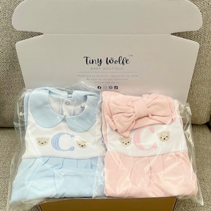 May include: A baby clothing set in a box. One set is light blue, the other is pink. Both have a white bib with the letter "C" and teddy bear embroidery. The box reads "Tiny Wolfe Baby Boutique".