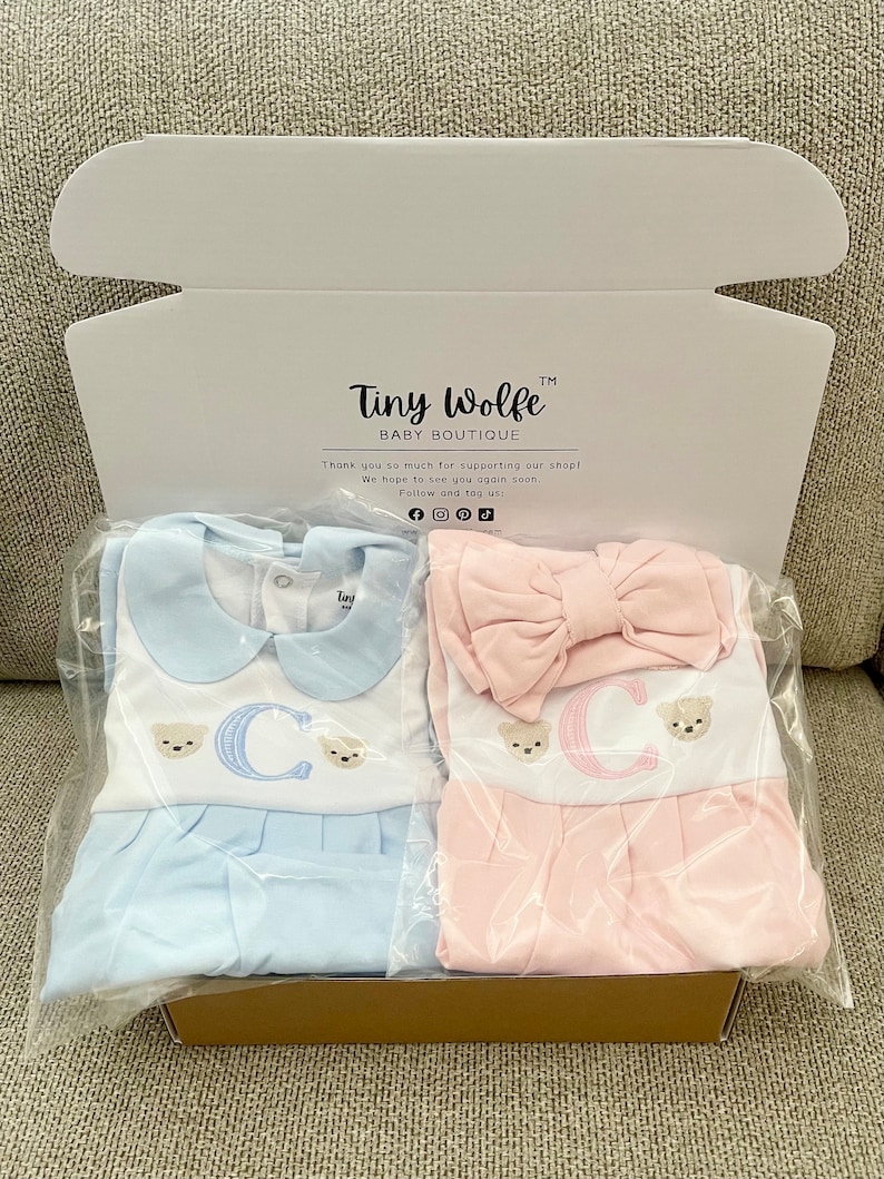 May include: A baby clothing set in a box. One set is light blue, the other is pink. Both have a white bib with the letter "C" and teddy bear embroidery. The box reads "Tiny Wolfe Baby Boutique".