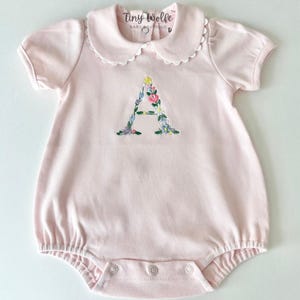 May include: A light pink baby romper with a white scalloped collar and short sleeves. The letter "A" is embroidered on the front with colorful flowers. The text "Tiny Wolfe BABY BOUTIQUE" is printed on the collar.