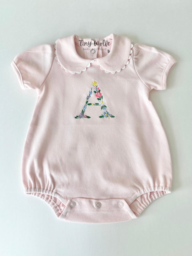May include: A light pink baby romper with a white scalloped collar and short sleeves. The letter "A" is embroidered on the front with colorful flowers. The text "Tiny Wolfe BABY BOUTIQUE" is printed on the collar.