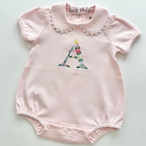 May include: A light pink baby romper with short sleeves and a Peter Pan collar. The letter "A" is embroidered with colorful flowers. The romper has snap closures and the text "Tiny Wolfe Baby Boutique" is visible.