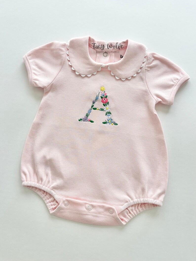 May include: A light pink baby romper with short sleeves and a Peter Pan collar. The letter "A" is embroidered with colorful flowers. The romper has snap closures and the text "Tiny Wolfe Baby Boutique" is visible.