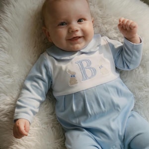 May include: A baby wearing a light blue and white romper with a Peter Pan collar. The romper features an embroidered letter "B" and two bunny designs. The baby is lying on a fluffy white surface.