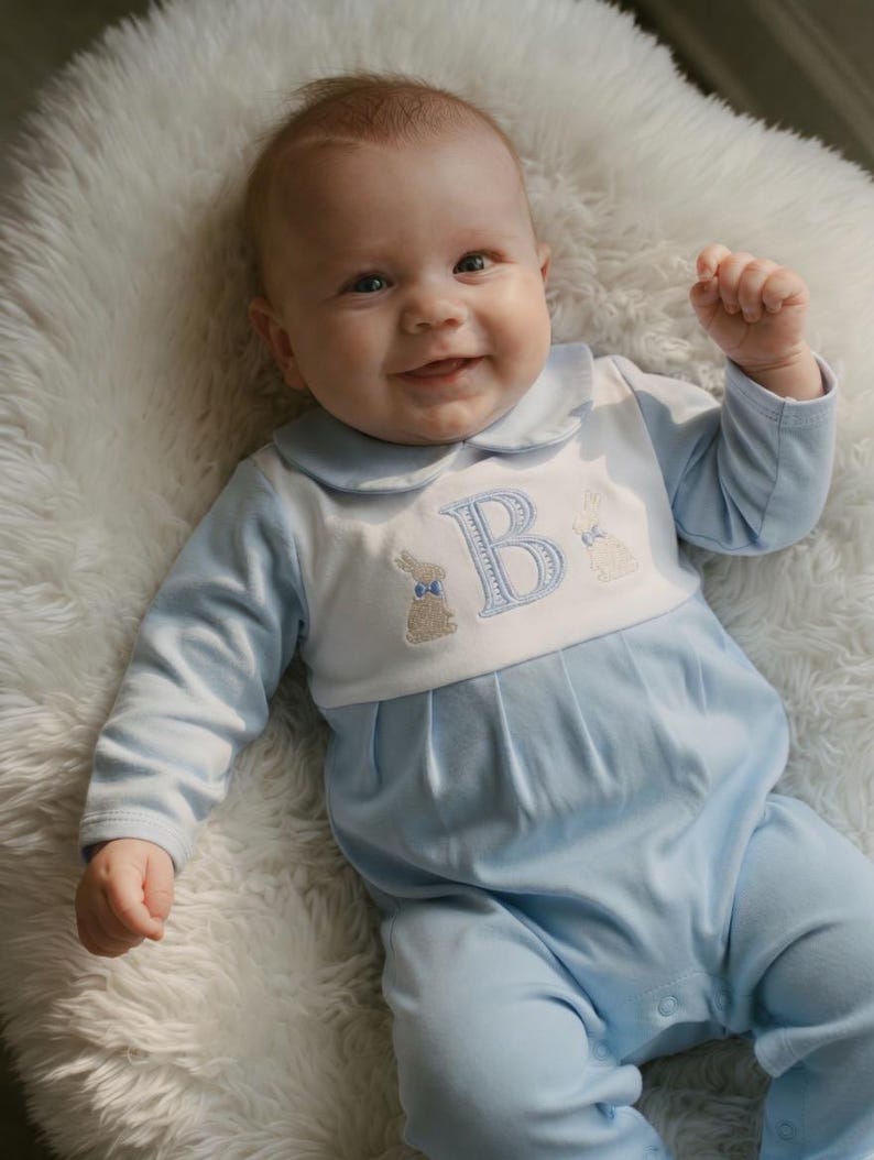 May include: A baby wearing a light blue and white romper with a Peter Pan collar. The romper features an embroidered letter "B" and two bunny designs. The baby is lying on a fluffy white surface.