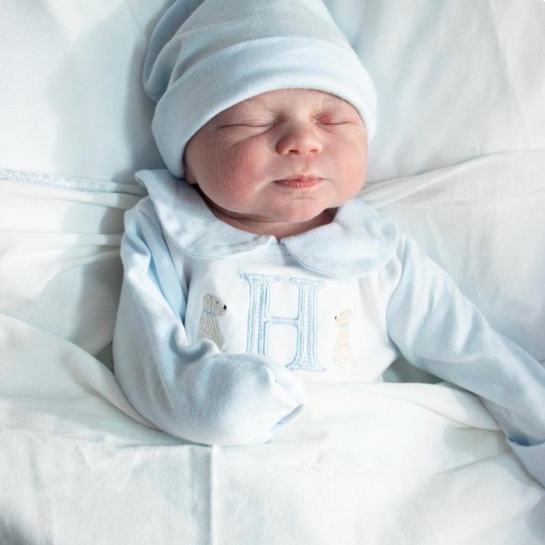 May include: A newborn baby wearing a light blue hat and a matching long-sleeved onesie. The onesie features a Peter Pan collar, embroidered teddy bears, and a large letter "H". The baby is lying on a white blanket.