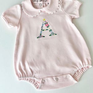 May include: A light pink baby romper with a Peter Pan collar and short sleeves. The romper features an embroidered floral "A" on the front. The brand name "Tiny Wolfe Baby Boutique" is printed on the collar.