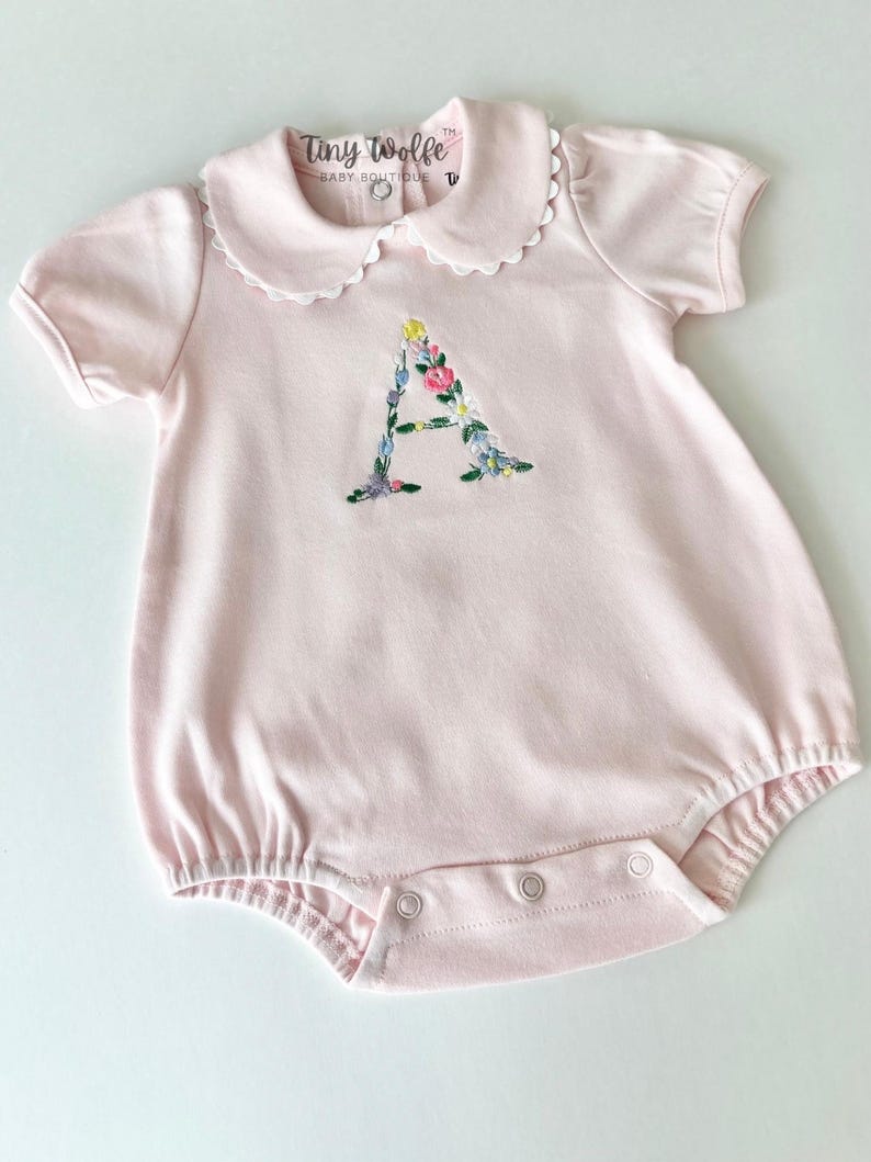 May include: A light pink baby romper with a Peter Pan collar and short sleeves. The romper features an embroidered floral "A" on the front. The brand name "Tiny Wolfe Baby Boutique" is printed on the collar.