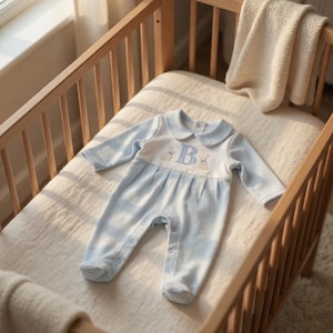 May include: A light blue baby romper with a white collar and the letter "B" embroidered on the chest. The romper features a striped pattern and is placed in a wooden crib with a cream-colored blanket.