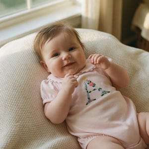 May include: A baby wearing a light pink onesie with a floral embroidered letter "A" on the front. The onesie has a Peter Pan collar and short sleeves. The baby is lying on a cream-colored blanket.