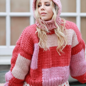 Könnte beinhalten: Ein gestrickter Pullover mit rosa und roten Streifen und einer passenden Strickmütze. Der Pullover hat eine grobmaschige Strickstruktur und eine lockere Passform.