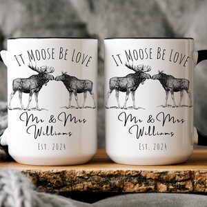 May include: Two white ceramic mugs with black handles and rims. Each mug features a black and white illustration of two moose and the text "IT MOOSE BE LOVE", "Mr & Mrs Williams", and "EST. 2024".
