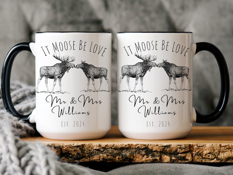 May include: Two white ceramic mugs with black handles and rims. Each mug features a black and white illustration of two moose and the text "IT MOOSE BE LOVE", "Mr & Mrs Williams", and "EST. 2024".