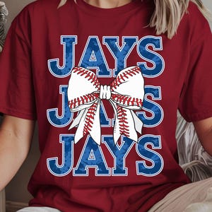 Könnte beinhalten: Burgunderfarbenes T-Shirt mit dem Wort "JAYS" in blauen Buchstaben mit Used-Look. Eine Baseball-Themen-Schleife mit weißen und roten Nähten ist mittig auf dem Shirt platziert. Ein lässiger, sportlicher Stil.