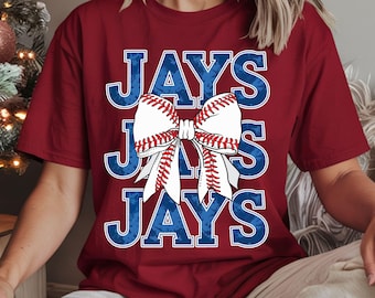 May include: Burgundy t-shirt featuring the word "JAYS" in blue letters with a distressed texture. A baseball-themed bow graphic is centered on the shirt, with white and red stitching details. The shirt is a casual, sporty style.