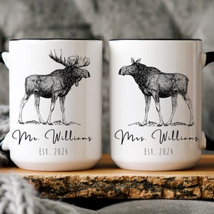 May include: Two white ceramic mugs with black handles and rims. Each mug features a detailed black and white illustration of a moose. One mug says "Mr. Williams EST. 2024" and the other says "Mrs. Williams EST. 2024".