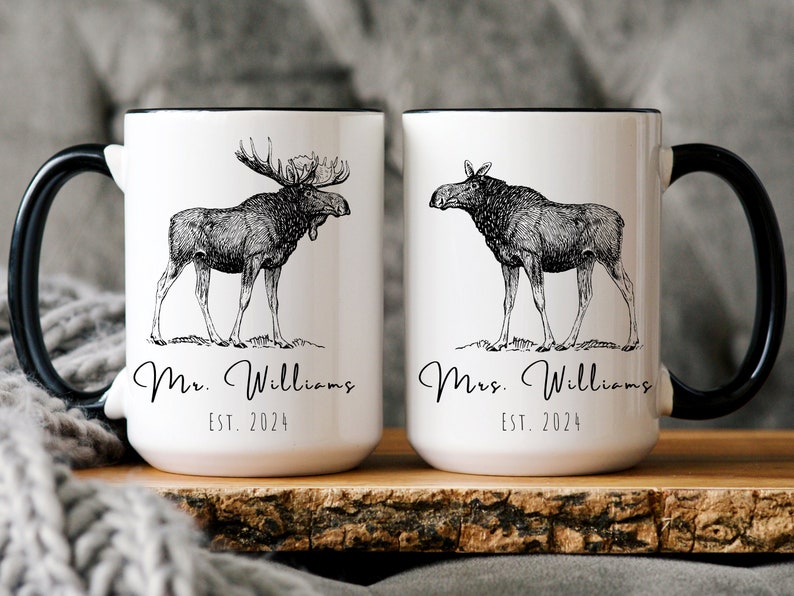 May include: Two white ceramic mugs with black handles and rims. Each mug features a detailed black and white illustration of a moose. One mug says "Mr. Williams EST. 2024" and the other says "Mrs. Williams EST. 2024".