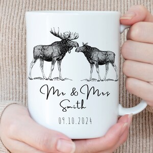 May include: White ceramic mug featuring a black and white illustration of two moose. The mug is personalized with the text "Mr & Mrs Smith" and the date "09.10.2024". The mug is held by a person.