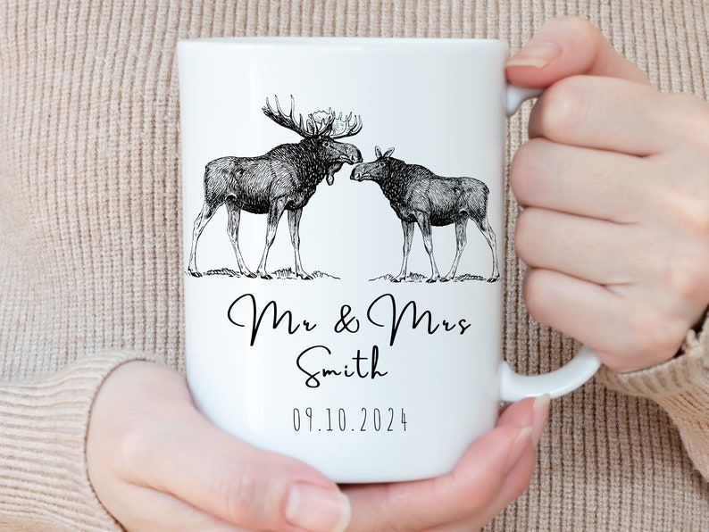 May include: White ceramic mug featuring a black and white illustration of two moose. The mug is personalized with the text "Mr & Mrs Smith" and the date "09.10.2024". The mug is held by a person.