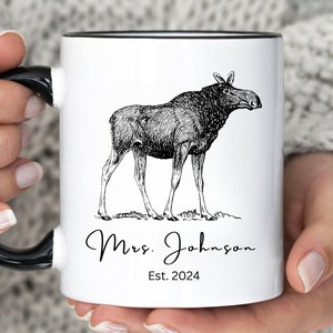 May include: White ceramic mug with a black handle and rim. The mug features a detailed black and white illustration of a moose. Below the moose is the text "Mrs. Johnson" and "Est. 2024" in a script font.