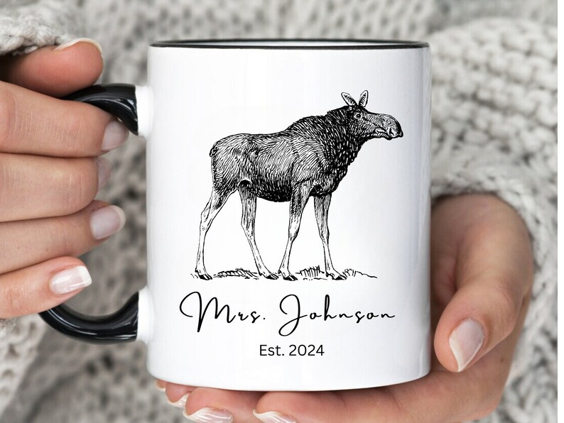 May include: White ceramic mug with a black handle and rim. The mug features a detailed black and white illustration of a moose. Below the moose is the text "Mrs. Johnson" and "Est. 2024" in a script font.