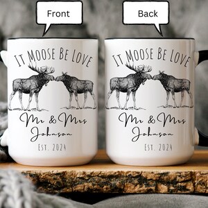 May include: White ceramic mug with black handle and rim, featuring a moose couple illustration and the text "IT MOOSE BE LOVE". Below the illustration is "Mr & Mrs Johnson EST. 2024".