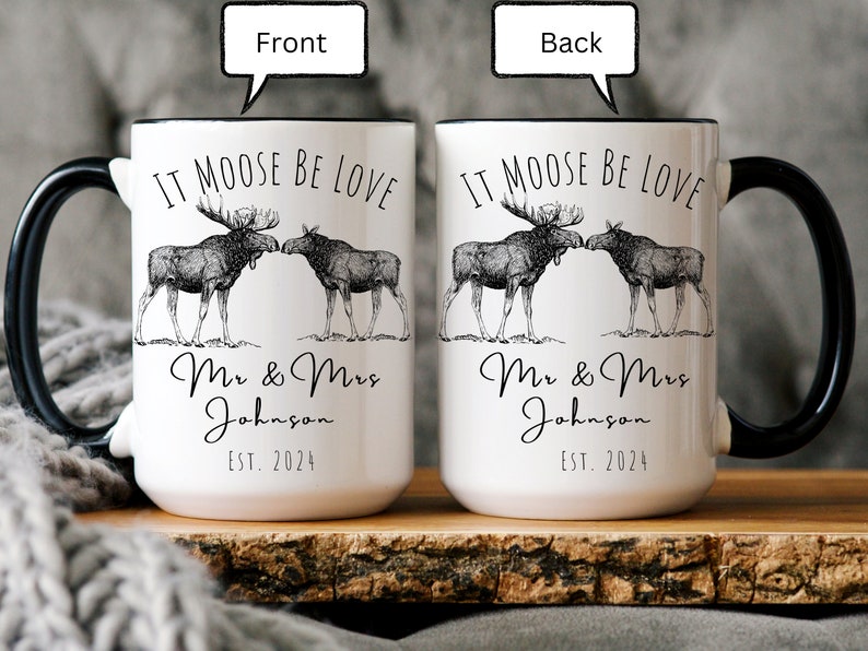 May include: White ceramic mug with black handle and rim, featuring a moose couple illustration and the text "IT MOOSE BE LOVE". Below the illustration is "Mr & Mrs Johnson EST. 2024".
