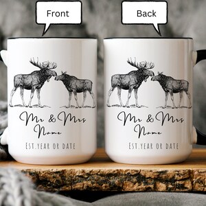 May include: White ceramic mug with a black handle and rim. The front and back of the mug feature a detailed illustration of two moose, with the text "Mr & Mrs Name" and "Est. Year or Date" below the moose. A rustic design.