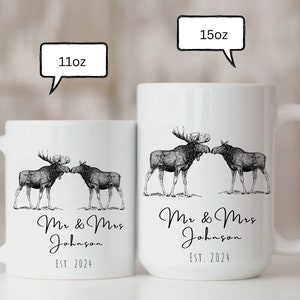 May include: White ceramic mugs with a black and white illustration of two moose touching noses. The mugs are labeled "11oz" and "15oz" and feature the text "Mr & Mrs Johnson EST. 2024".