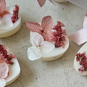 Può includere: Tavolette di cera bianca rotonde decorate con petali di ortensia rosa essiccati, foglie scheletrizzate bianche e piccoli grappoli di fiori bordeaux essiccati. Ogni tavoletta presenta una superficie liscia e un nastro rosa tenue.