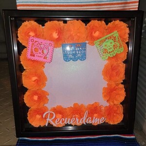 May include: A framed shadow box with orange paper flowers arranged around the edges. The box has a white background with the words "Recuérdame" written in white. There are also three paper cutouts with skull designs in pink, blue, and green.