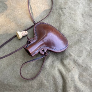 May include: A brown leather flask with a cork stopper and a brown leather strap. The flask is lying on a green canvas background.