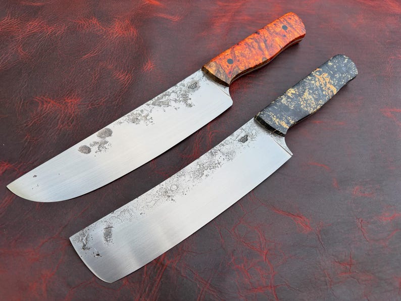 Pu&ograve; includere: Due coltelli da cucina fatti a mano con lame rustiche e usurate. Un coltello ha un manico con venature arancioni e marroni, l'altro ha un manico nero e dorato. Le lame sono argentate e mostrano segni di usura.