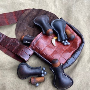 May include: A brown leather pouch with a crocodile pattern, containing several small black leather pouches with wooden stoppers and metal balls. The pouch is also decorated with a bone-like object.