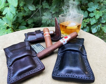 May include: Four leather cigar cases, two brown and two black, with a cigar and a lighter on top of them. A glass of amber liquid with ice and steam rising from it is in the background.