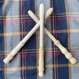 May include: Three wooden dowels, each with a rounded end, are arranged in an X shape on a plaid fabric background. The dowels are light brown in color and have a smooth, polished finish.