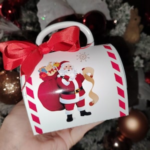 May include: White ceramic mug with a red ribbon and a Santa Claus design. The mug has a red and white striped pattern on the side. The Santa Claus is carrying a bag of gifts and a scroll.