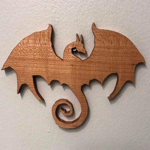 May include: A wooden dragon wall hanging with outstretched wings and a curled tail. The dragon is cut from light brown wood and has a detailed design. The dragon is mounted on a white wall.