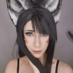 May include: A woman with long black hair and gray wolf ears is wearing brown eyeliner with a red and white design. She is wearing a black tank top.