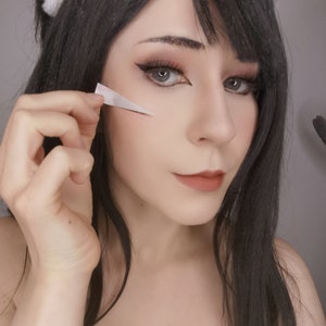May include: A woman with long black hair and cat ears is applying a white strip of tape to her face. She is wearing black eyeliner and lipstick.
