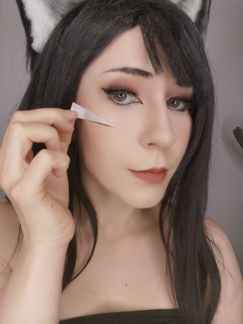 May include: A woman with long black hair and cat ears is applying a white strip of tape to her face. She is wearing black eyeliner and lipstick.