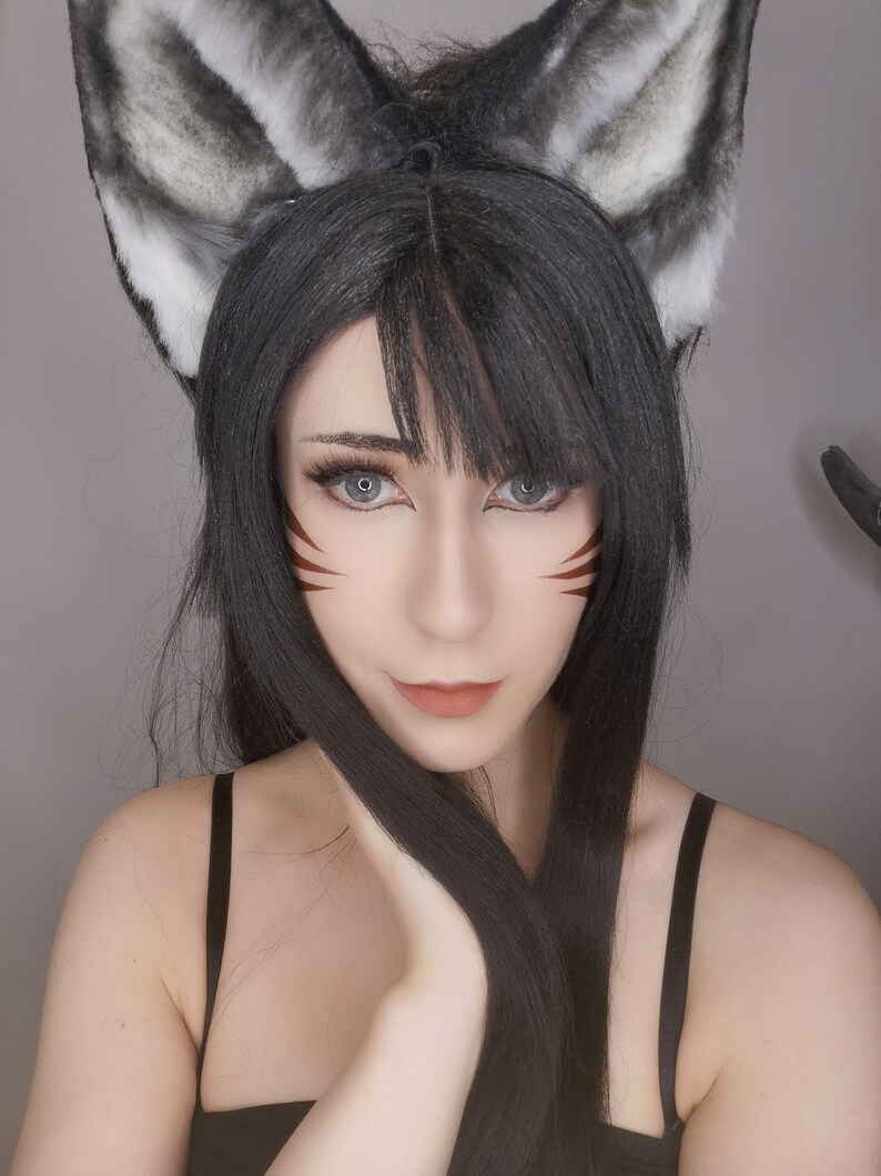May include: A woman with long black hair and gray wolf ears is wearing brown eyeliner with a red and white design. She is wearing a black tank top.