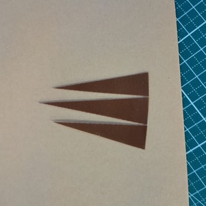 May include: Three brown leather triangles cut out of a larger piece of leather. The leather is a light brown color and the triangles are a darker brown color.