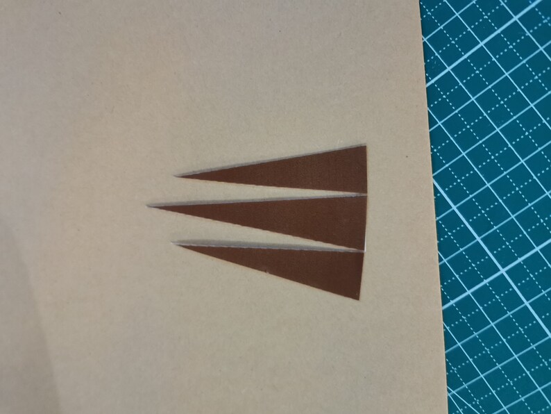 May include: Three brown leather triangles cut out of a larger piece of leather. The leather is a light brown color and the triangles are a darker brown color.