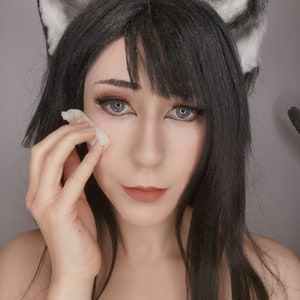 May include: A woman with long black hair wearing cat ears is applying makeup with a cotton pad.