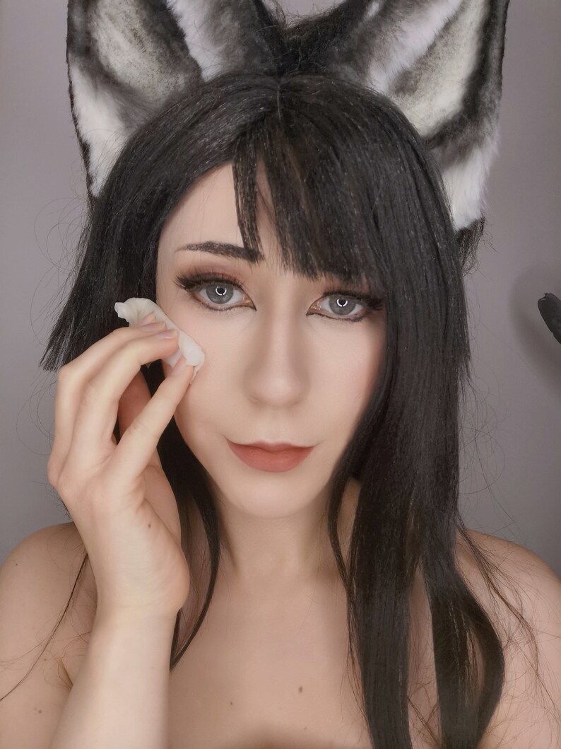 May include: A woman with long black hair wearing cat ears is applying makeup with a cotton pad.