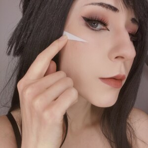 May include: A woman with long black hair is wearing makeup, including white face paint on her cheek. She is touching the paint with her finger.