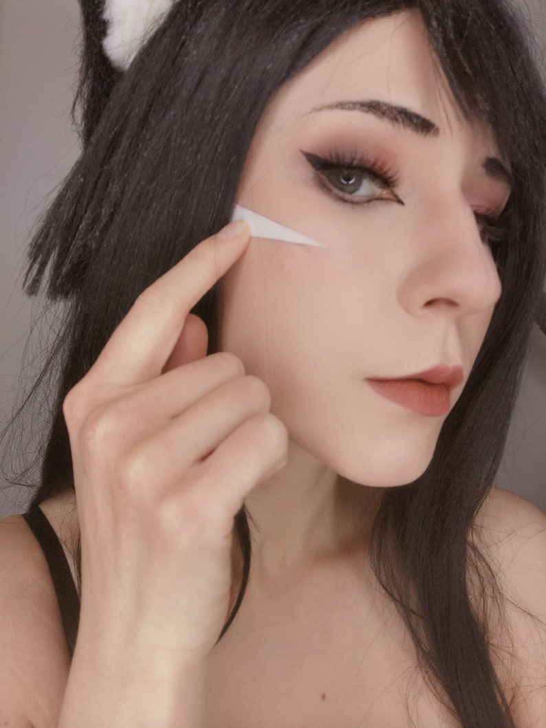 May include: A woman with long black hair is wearing makeup, including white face paint on her cheek. She is touching the paint with her finger.