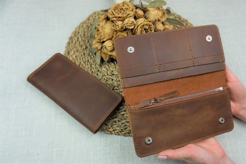 Puede incluir: Una cartera de cuero marr&oacute;n con m&uacute;ltiples ranuras para tarjetas y un compartimento con cremallera. La cartera est&aacute; abierta y el interior es visible.