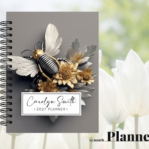 May include: A gray planner with a silver and gold bee and flowers on the cover. The planner is titled "Carolyn Smith 2027 Planner".